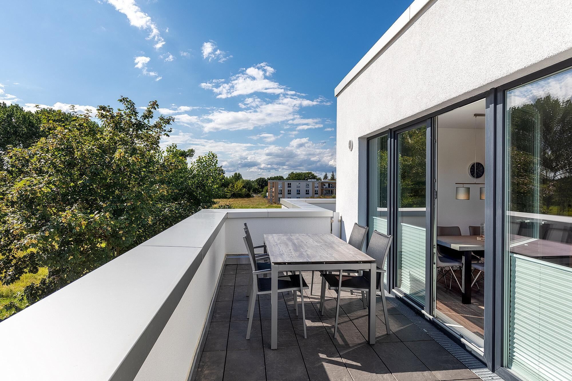 Um pátio externo moderno com uma mesa e cadeiras, grandes portas de vidro deslizantes e vista para um edifício e árvores ao fundo.