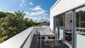 A modern outdoor patio with a table and chairs, large sliding glass doors, and a view of a building and trees in the background. - Strandnahes Familienapartment mit Toller, Sonniger Dachterrasse und Zwei Bädern (Ostseebad Boltenhagen)
