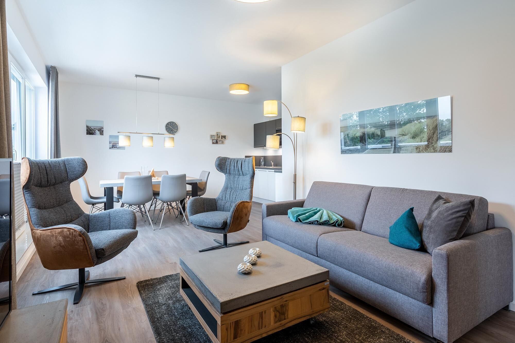 Ein modernes Wohnzimmer mit grauem Sofa, zwei Sesseln, einem Couchtisch und einem Essbereich mit Tisch und Stühlen.