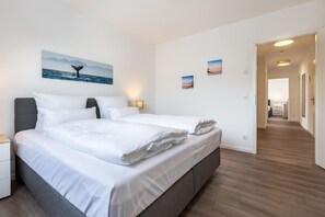A hotel room with a large bed, a nightstand, a desk, and a desk lamp. There are three wall-mounted pictures and a door leading to another room.