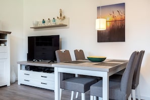 Un comedor moderno con una mesa de madera, sillas tapizadas de color gris, un bol verde y un televisor sobre un mueble blanco.