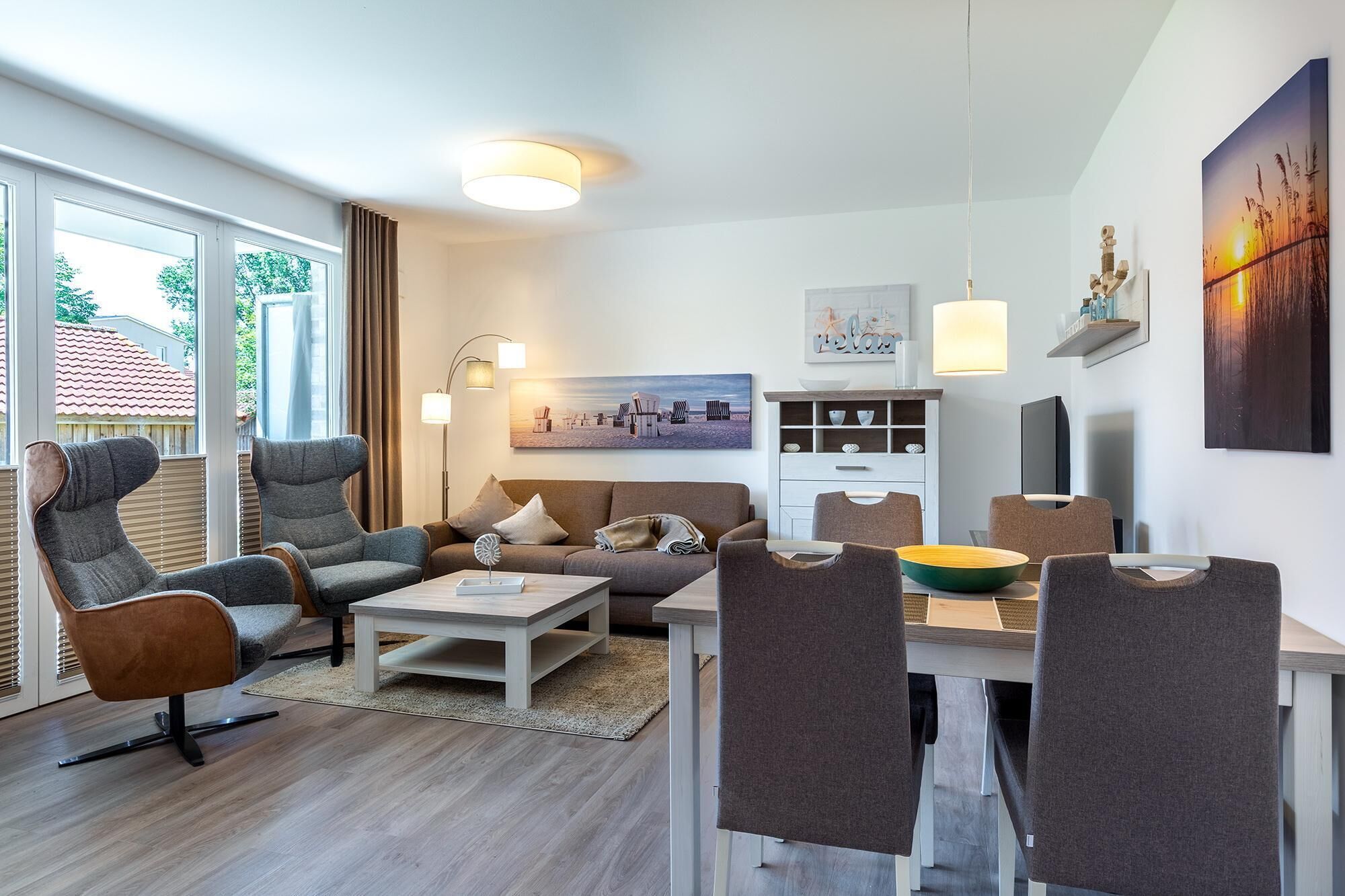 Una sala moderna con un sofá, sillones, una mesa de centro y un comedor con sillas y una mesa. Hay un cuadro en la pared, una lámpara y un televisor sobre un mueble.