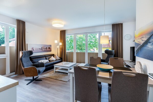 A modern living room with a sofa, chairs, a coffee table, and a dining table. Large windows with curtains provide natural light and views of greenery outside. - Liebevoll Eingerichtetes Apartment mit Tollem, Sonnigem Balkon in Strandnähe (Ostseebad Boltenhagen)