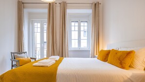 Ein ordentlich bezogenes Bett mit gelber Decke und Kissen, ein Stuhl mit Sitzkissen und ein Fenster mit Vorhängen.