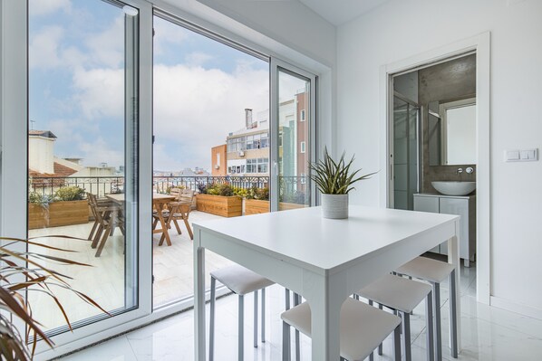A modern kitchen with a large window, a dining area with a white table and chairs, and a potted plant on the table.