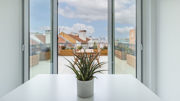 Ein Balkon mit einer Topfpflanze, einem Tisch und Blick auf die Stadt durch offene Schiebetüren.