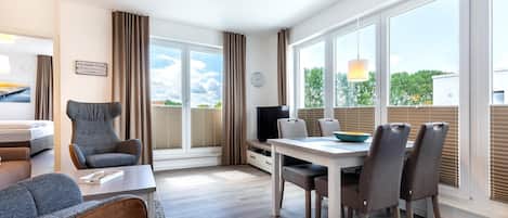A modern hotel room with a dining area, a sofa, a TV, and large windows with blinds.