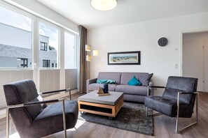 Una sala de estar moderna con un sofá gris, dos sillones, una mesa de centro y un amplio ventanal con persianas.