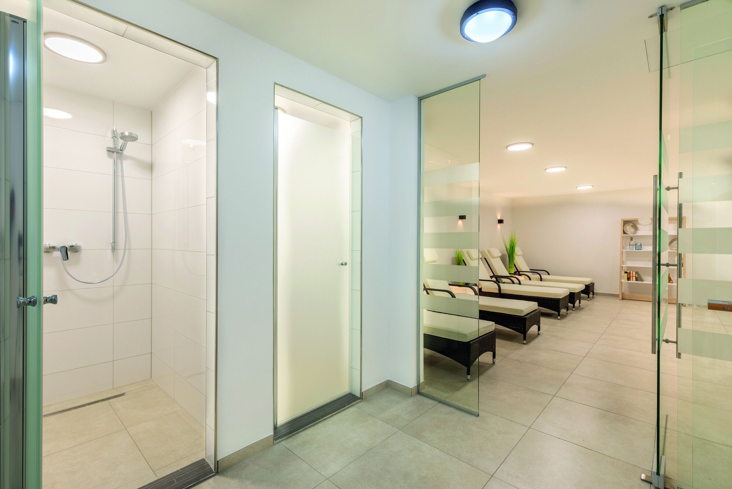 Une salle de bain moderne dotée d’une douche à parois de verre, d’une porte en verre et d’un coin salon avec des chaises et une plante.