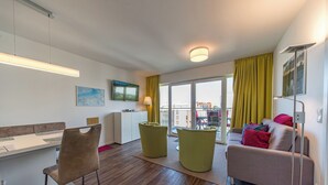 A modern living room with a sofa, two green ottomans, a dining table with chairs, a flat-screen TV, and a large window with curtains. - Great apartment with infrared sauna and balcony with a wonderful panoramic view! (Nordseeheilbad Schillig)