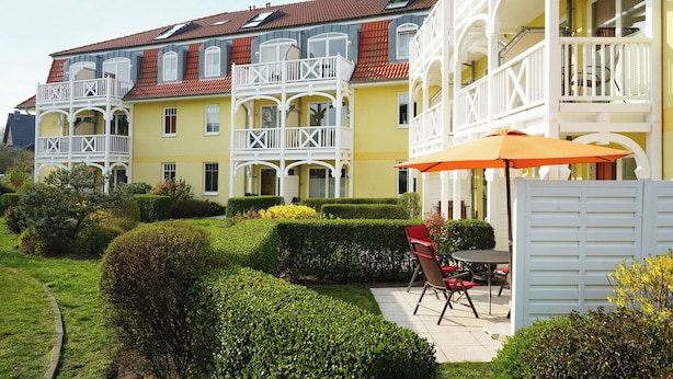 Eine Reihe von gelb-weißen Gebäuden mit Balkonen, ein Garten mit Hecken und ein Tisch mit Stühlen unter einem Sonnenschirm.