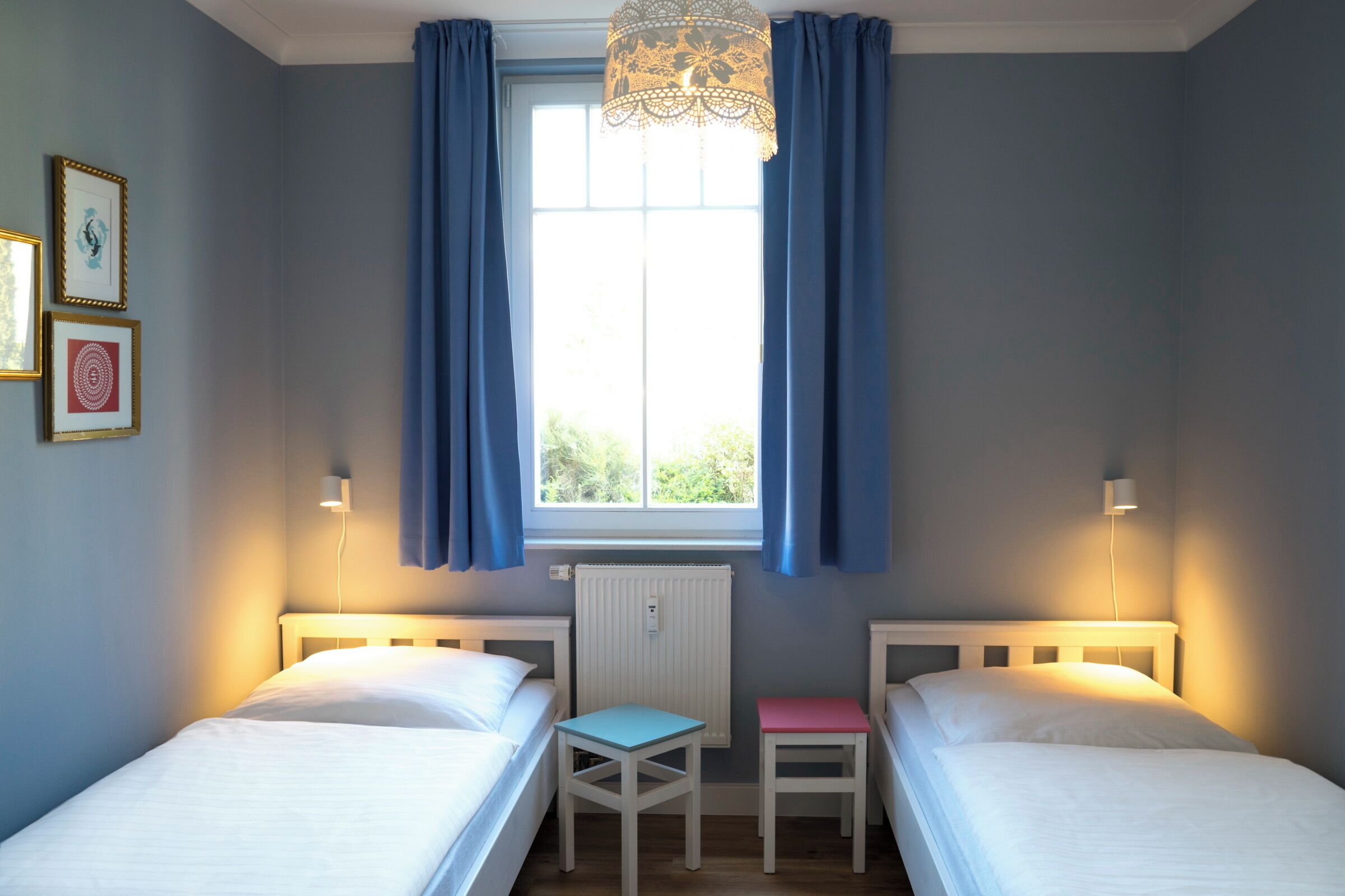 A bedroom with two beds, a window with curtains, a radiator, and a table.