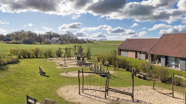 Un paisaje rural con un parque infantil, un edificio de techo rojo y un cielo despejado.