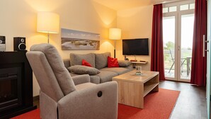Un salón moderno con un sofá gris, cojines rojos, una mesa de centro de madera y un televisor de pantalla plana. Hay una chimenea, un cuadro en la pared y vistas a través de la puerta de cristal.