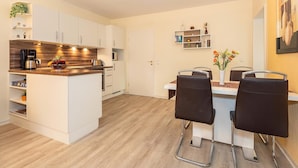 Una cocina moderna con armarios blancos, una cafetera y un frutero. Hay una zona de comedor con una mesa blanca y sillas negras.
