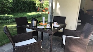Un patio con una mesa dispuesta para cuatro personas, rodeado de sillas de mimbre, una cafetera y un florero con flores.