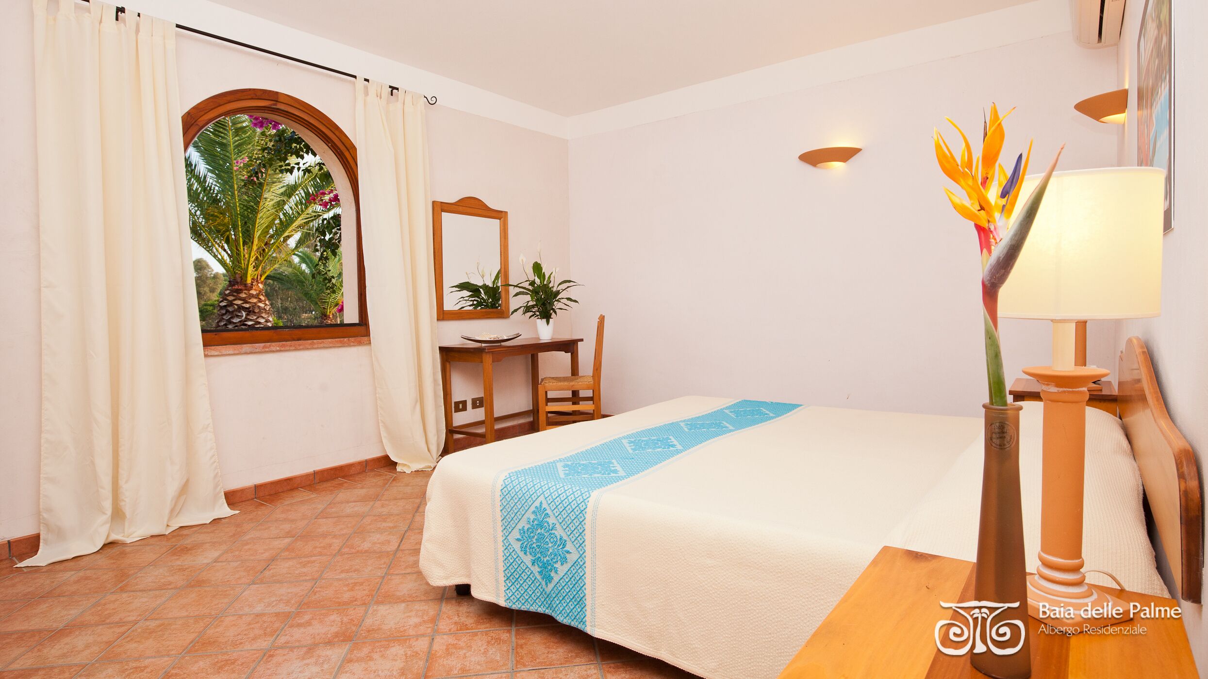 A hotel room with a bed, a desk, a chair, a window with curtains, a mirror, a plant, and a lamp.
