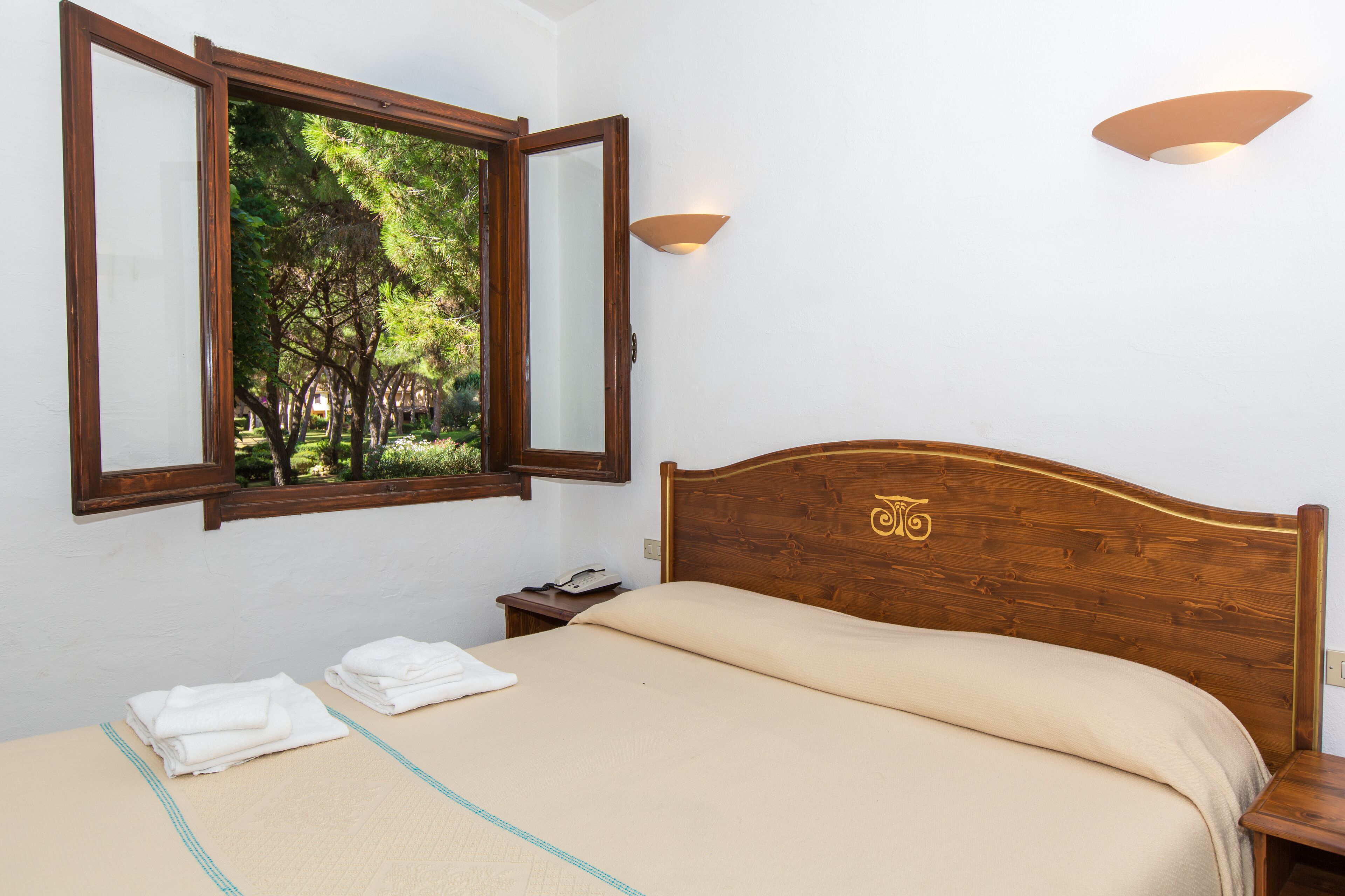 A bedroom with a wooden headboard, a bed with a beige bedspread, a window with a view of trees, and two wall-mounted lights.