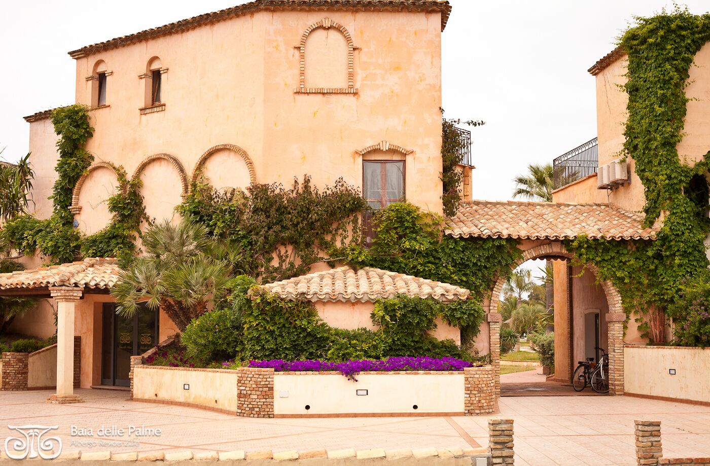 Un bâtiment doté de fenêtres en arc de cercle, d’un toit recouvert de tuiles et d’un jardin où poussent des fleurs violettes.