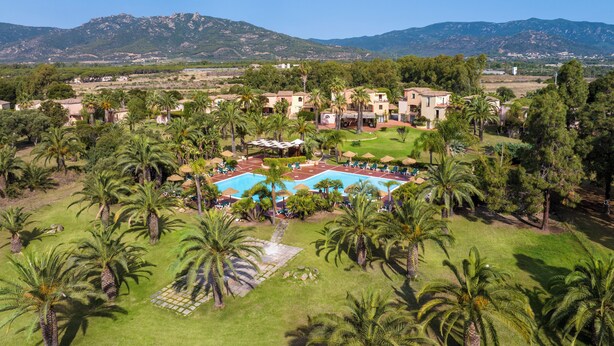 A resort with a swimming pool surrounded by palm trees and a mountainous backdrop. - Residence Baia delle Palme (Pula)