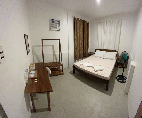 A hotel room with a bed, a desk, a chair, a bedside table, a fan, and a window with curtains. - Hill Myna Beach Cottages (Puerto Princesa)