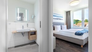 Un baño moderno con un espejo grande, un lavamanos blanco y almacenamiento de madera. El dormitorio tiene una cama grande, un cabecero gris y vistas a la vegetación a través de un ventanal amplio.