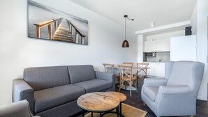 Una sala de estar moderna con un sofá gris, una mesa de centro redonda de madera y un sillón gris. En el fondo, se ve una zona de comedor con una mesa y sillas de madera.