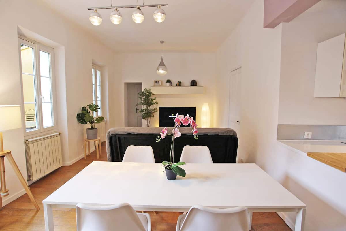Un espace de salle à manger moderne avec une table blanche, des chaises et une orchidée en pot servant de centre de table. Un luminaire suspendu se trouve au-dessus, et une plante est posée sur un petit support à côté de la table.