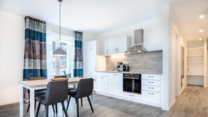 Una cocina moderna con armarios blancos, horno integrado y cafetera. Hay una mesa de comedor con dos sillas y una lámpara colgante.