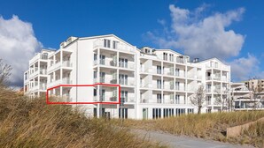 Un edificio blanco de varios pisos con balcones, situado cerca de una zona arenosa con vegetación.