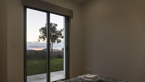 Ein Schlafzimmer mit einer großen Glasschiebetür, die auf einen Balkon führt, einem Bett mit grauer Decke und einem kleinen schwarzen Korb.