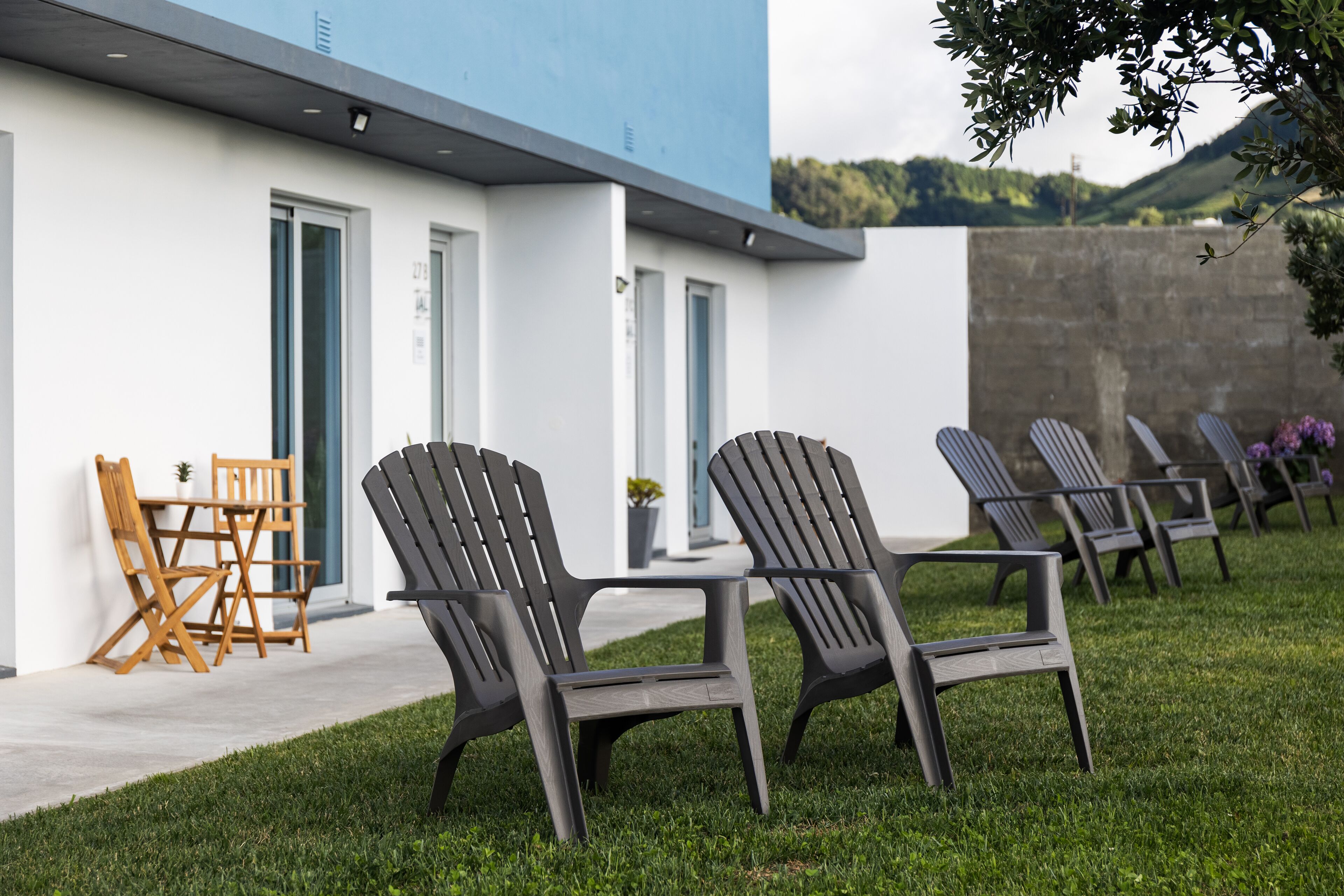 A modern outdoor seating area with Adirondack chairs and a wooden table, adjacent to a building with large windows and a blue facade.