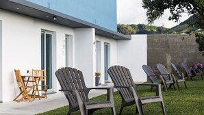 Ein moderner Außenbereich mit Adirondack-Stühlen und einem Holztisch, neben einem Gebäude mit großen Fenstern und einer blauen Fassade.