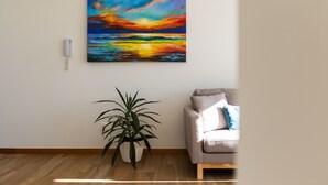 Ein Zimmer mit Holzboden, einem beigen Sofa, einer Zimmerpflanze und einem Gemälde, das einen Sonnenuntergang über dem Meer zeigt.