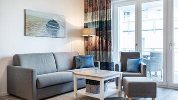 Ein modernes Wohnzimmer mit einem grauen Sofa, einem kleinen Tisch und einem grauen Sessel. Ein großes Fenster bietet Blick auf Gebäude, und ein Vorhang mit Blumenmuster ist angebracht.