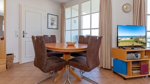 A dining area with a round table, four brown chairs, a television on a wooden stand, and a window with curtains.