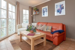 A living room with a white sofa, an orange sofa, a wooden coffee table, and a floor lamp. There are large windows, a bookshelf, and two framed pictures on the wall. - Helles Ostsee-apartment mit Balkon und Direktem Meerblick in Erstklassiger Lage! (Ostseebad Kühlungsborn)