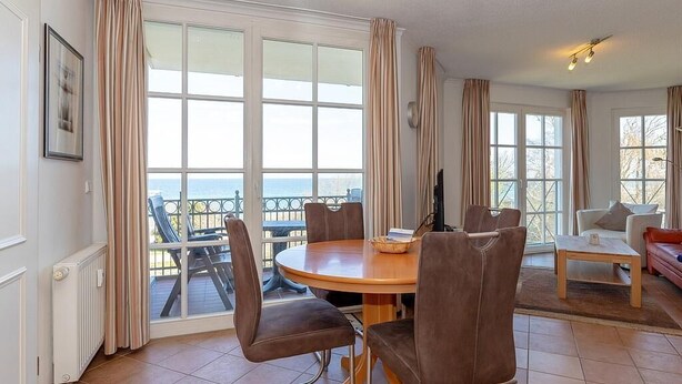 A dining area with a round table and chairs, large windows with curtains, and a view of the sea.