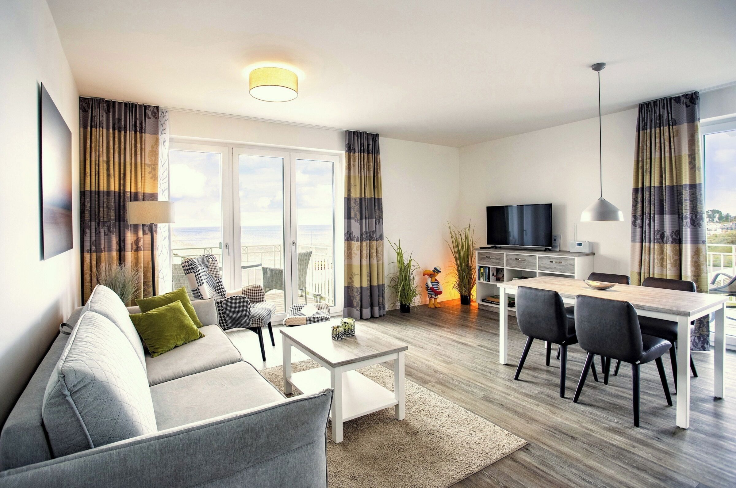 A modern living room with a sofa, coffee table, dining table, and chairs. There is a TV, a lamp, and a view of the outdoors through large windows.