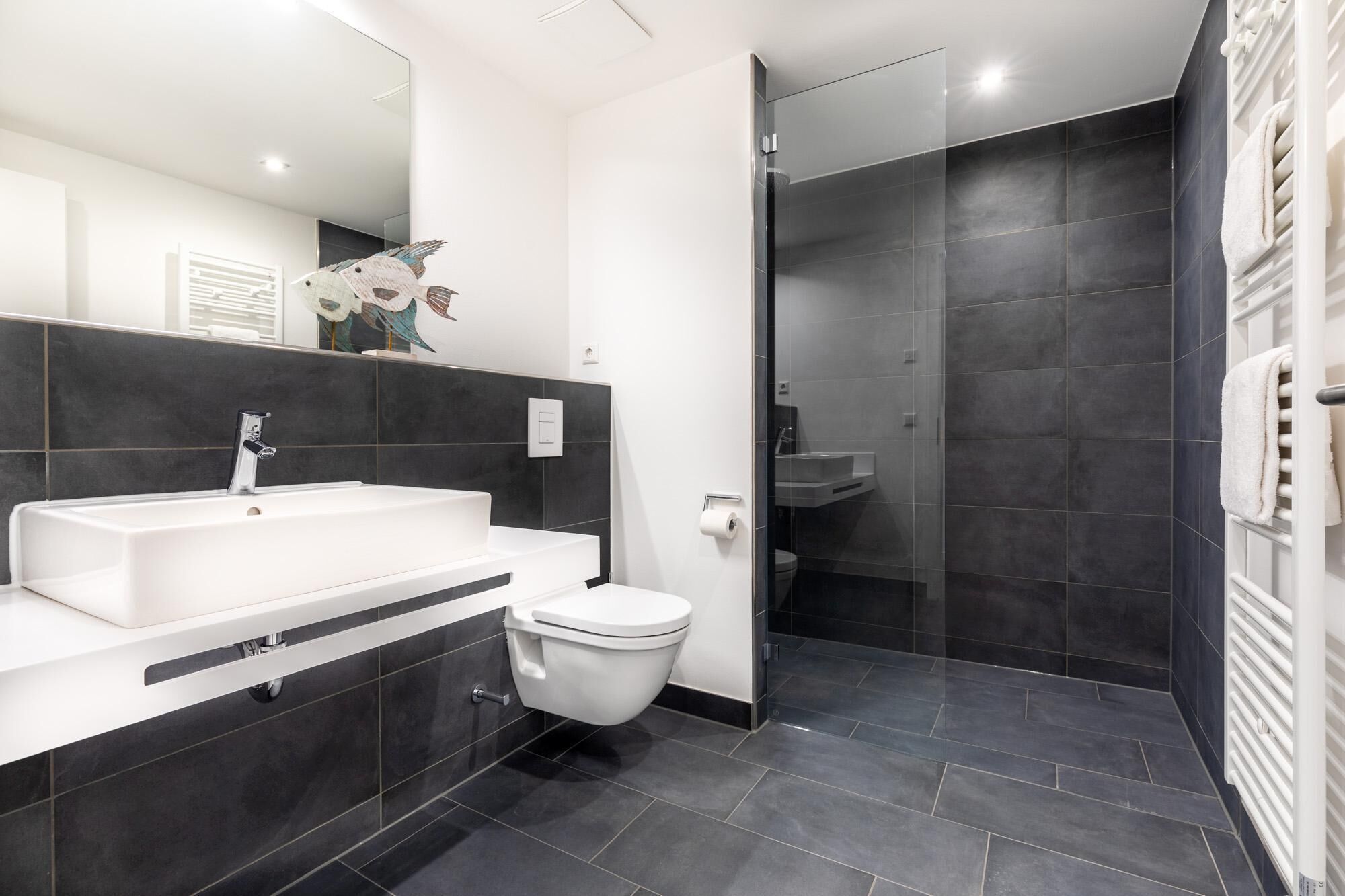 Un baño moderno con un lavamanos blanco, paredes cubiertas de baldosas oscuras, un inodoro y una cabina de ducha de vidrio.