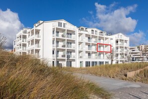 Un edificio de apartamentos blancos de varios pisos con balcones, cerca de una playa con pasto seco y un camino de hormigón.