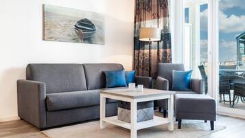 Ein modernes Wohnzimmer mit einem grauen Sofa, zwei Sesseln, einem Couchtisch und einem großen Fenster mit Blick auf den Strand.