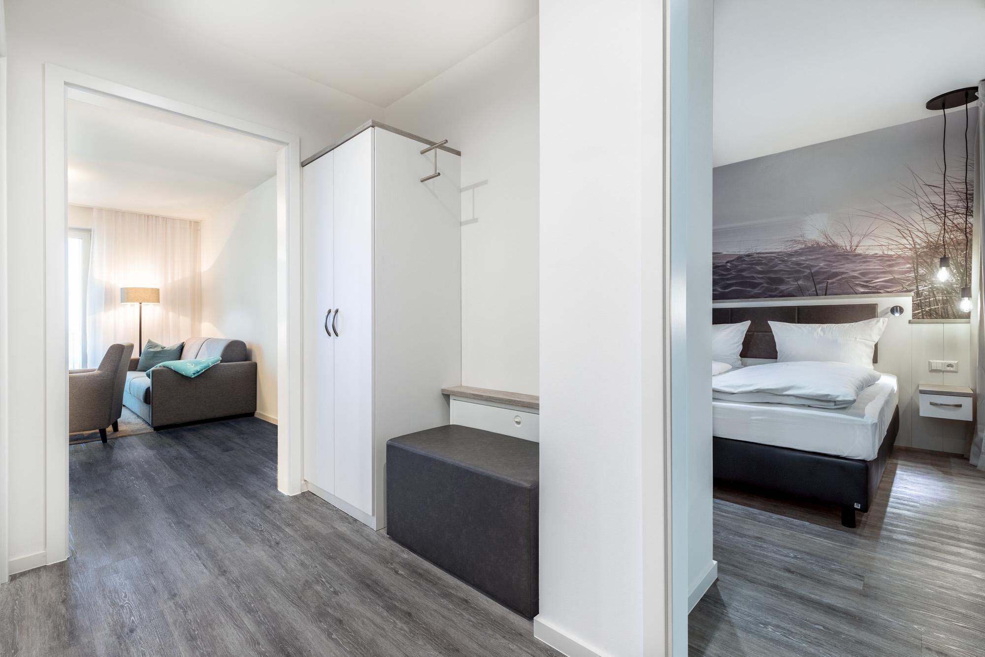 Une chambre d’hôtel moderne avec un plancher en bois gris, un canapé gris, un lit avec une literie blanche, une armoire blanche et un tableau représentant un paysage désertique accroché au mur.