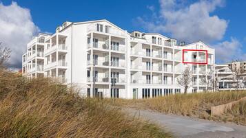 Ein mehrstöckiges weißes Gebäude mit Balkonen, in der Nähe einer sandigen Fläche mit Vegetation.