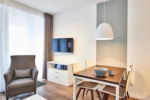 Una habitación de hotel moderna con una mesa de comedor de madera, un sillón gris, un escritorio blanco y un televisor montado en la pared.