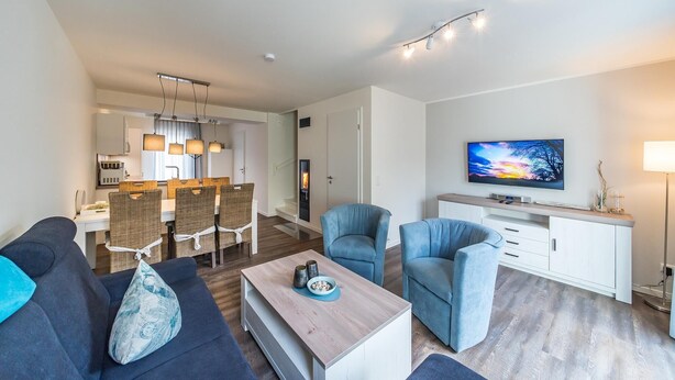 Un salon moderne avec un canapé, deux fauteuils, une table basse et un téléviseur. On y trouve également un coin repas avec une table et des chaises, et une cuisine à l’arrière-plan.