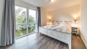 Une chambre avec une grande fenêtre, un lit recouvert de draps blancs, deux lampes de chevet et une table de chevet avec un tiroir.