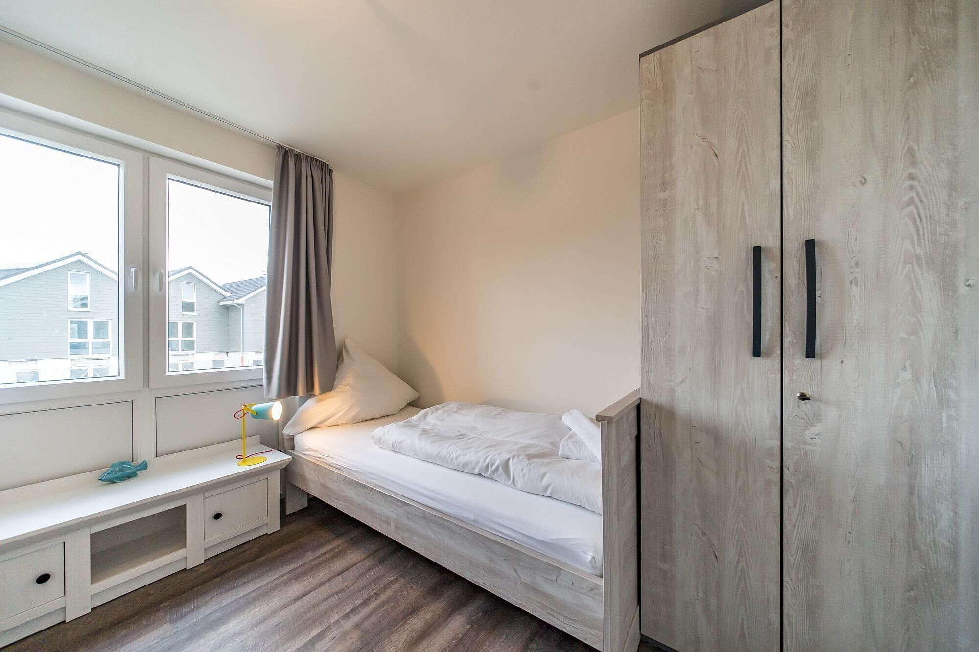 A bedroom with a bed, a nightstand, a lamp, a wardrobe, and a window with curtains.