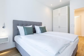A neatly made bed with white linens and two pillows, a grey upholstered headboard, and a white wardrobe with louvered doors.