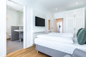 A modern bedroom with a grey sofa, a large bed with white bedding, a flat-screen TV mounted on the wall, and a walk-in closet with sliding doors.